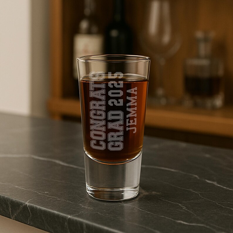 May include: A clear shot glass filled with a dark amber liquid. The glass has the words "CONGRATS GRAD 2025 JEMMA" etched in a bold, collegiate font. The glass sits on a dark, textured surface, with a blurred background.