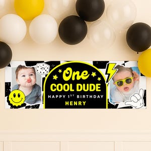 May include: A birthday banner with the text "One COOL DUDE Happy 1st Birthday HENRY" in yellow and white on a black background. The banner has two photos of a baby and is decorated with a smiley face, dice, and a lightning bolt. Balloons in yellow, white, and black are in the background.
