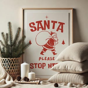 May include: A white framed poster with a red illustration of Santa Claus carrying a bag of gifts. The text on the poster reads "SANTA PLEASE STOP HERE".
