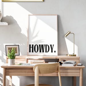 Op de afbeelding: Een zwart-witte print met het woord "HOWDY" in vetgedrukte, zwarte letters. De print is ingelijst en hangt aan een witte muur boven een houten bureau met boeken, een lamp en een stoel.