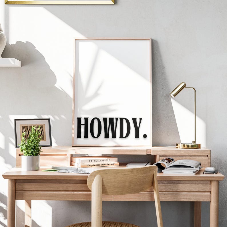 Op de afbeelding: Een zwart-witte print met het woord "HOWDY" in vetgedrukte, zwarte letters. De print is ingelijst en hangt aan een witte muur boven een houten bureau met boeken, een lamp en een stoel.