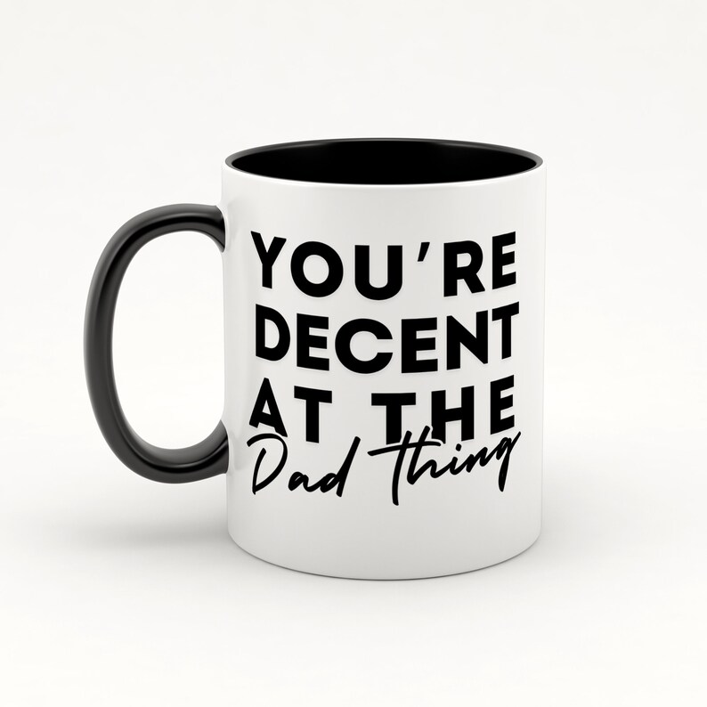May include: White ceramic coffee mug with a black handle and black rim. The mug has black text that reads "You're Decent at the Dad Thing."