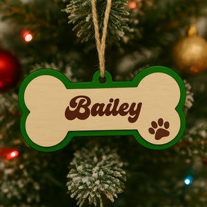 Pode incluir: Um enfeite de Natal em forma de osso de c&atilde;o de madeira com o nome "Bailey" em escrita castanha. O enfeite tem uma borda verde e uma pegada. Ele pende de uma corda de barbante natural.