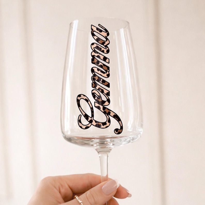May include: A clear champagne flute with the name "Emma" in a leopard print design. The stemware is held by a person with a ring on their finger. The glass is elegant and suitable for special occasions.