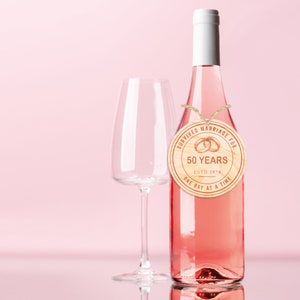 May include: A wooden round tag with the text "Survived Marriage For 50 Years Est. 1974 One Day At A Time" and two gold rings, tied with twine around the neck of a bottle of pink wine.