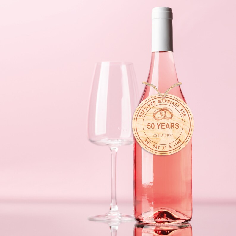 May include: A wooden round tag with the text "Survived Marriage For 50 Years Est. 1974 One Day At A Time" and two gold rings, tied with twine around the neck of a bottle of pink wine.