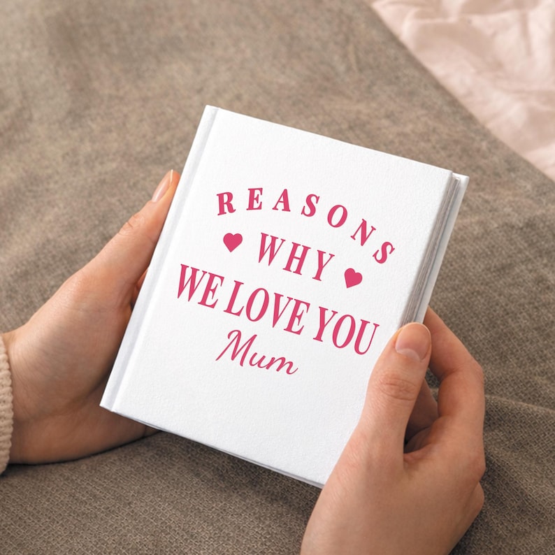 May include: A white book with the text "REASONS WHY WE LOVE YOU Mum" in pink script. Two pink hearts are placed before and after the word "WHY". The book is held open by a person.