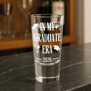May include: A clear shot glass with the text "IN MY GRADUATE ERA 2026" in white. The glass is filled with a clear liquid and sits on a dark surface. A graduation cap graphic is included.