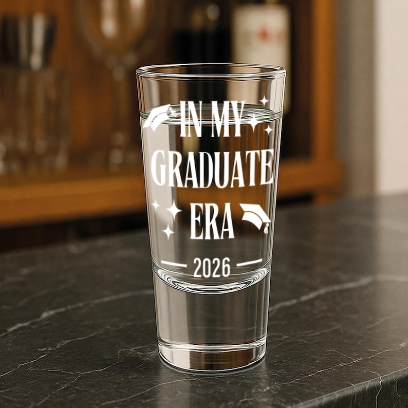 May include: A clear shot glass with the text "IN MY GRADUATE ERA 2026" in white. The glass is filled with a clear liquid and sits on a dark surface. A graduation cap graphic is included.