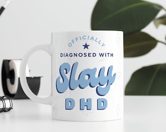 Pode incluir: Caneca de cerâmica branca com um design azul e branco. A caneca diz "Officially Diagnosed With Slay DHD" com uma estrela e raios.
