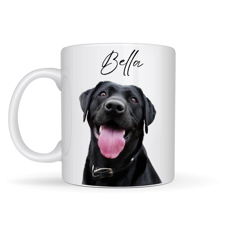 May include: White ceramic mug featuring a black Labrador Retriever with its tongue out. The dog is wearing a black collar. The name "Bella" is written in cursive above the dog's head.