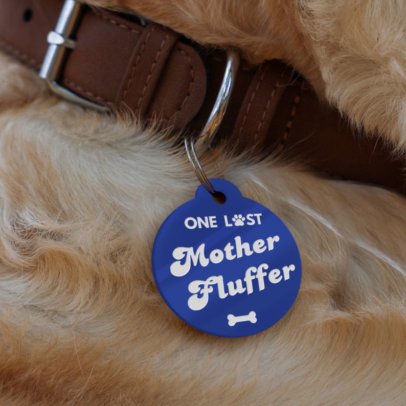 Op de afbeelding: Blauwe hondenhanger met witte tekst die "ONE LOST Mother Fluffer" luidt, met een bot-icoon onder de tekst.