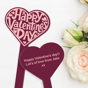 May include: Two burgundy heart-shaped signs with pink lettering. The top sign says "Happy Valentine's Day". The bottom sign says "Happy Valentine's day!! Lot's of love from Jake xx".