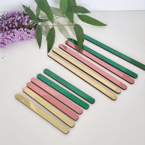 May include: A set of eight wooden craft sticks with glitter finishes. The sticks are arranged in a rainbow pattern, with gold, pink, and green glitter.