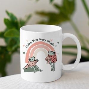 Puede incluir: Taza de cer&aacute;mica blanca con un arco&iacute;ris y dos ranas que llevan jers&eacute;is. El texto en la taza dice "I Like You Very Mush".