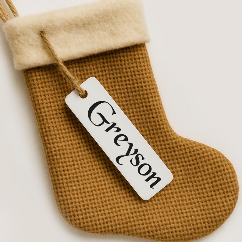 May include: A brown Christmas stocking with a textured waffle weave pattern. The stocking has a cream-colored cuff and a white tag with the name "Greyson" printed in black.