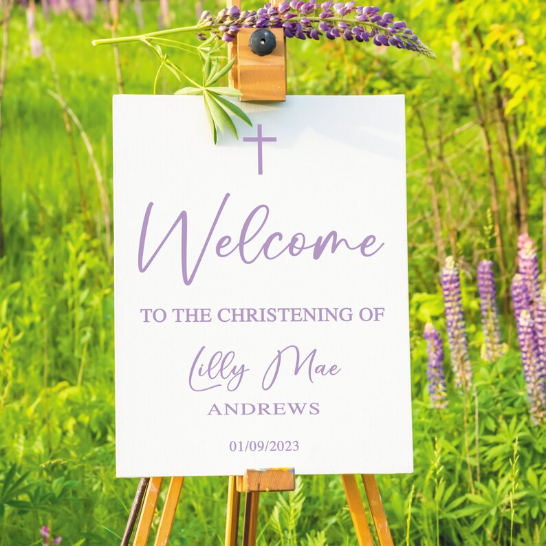 以下が含まれることがあります： 紫色の十字架と「Lilly Mae Andrews 01/09/2023 の洗礼式へようこそ」というテキストが書かれた白い看板が、紫の花が咲く畑の中にイーゼルに立てかけられています。