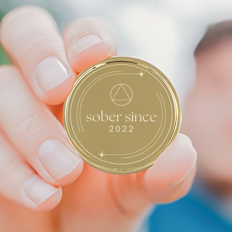 Puede incluir: Una moneda de color dorado con un s&iacute;mbolo de tri&aacute;ngulo y el texto "sobrio desde 2022" en ella.