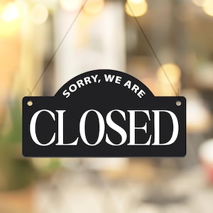 May include: A black rectangular sign with rounded top corners, hanging by a thin string. The sign reads "SORRY, WE ARE" above "CLOSED" in large white capital letters. The background is blurred.