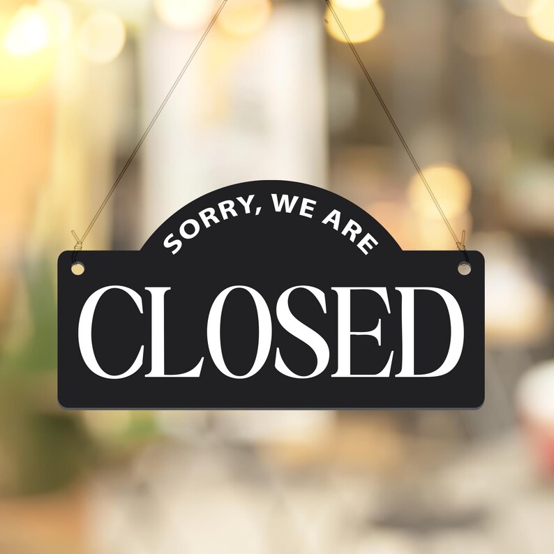 May include: A black rectangular sign with rounded top corners, hanging by a thin string. The sign reads "SORRY, WE ARE" above "CLOSED" in large white capital letters. The background is blurred.