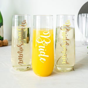 May include: Three champagne flutes with gold lettering. The flutes are filled with different beverages, including orange juice and champagne. The lettering on the flutes reads "Bride" and "Bridesmaid".