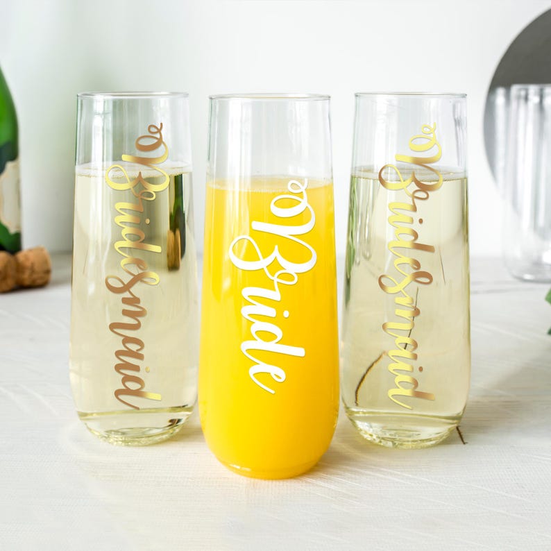 May include: Three champagne flutes with gold lettering. The flutes are filled with different beverages, including orange juice and champagne. The lettering on the flutes reads "Bride" and "Bridesmaid".
