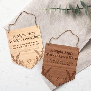 May include: Two wooden signs with rope hangers. The signs read "A Night Shift Worker Lives Here" with instructions not to knock or ring the doorbell. Floral and heart designs are etched on the signs. One sign is light wood, the other is dark.