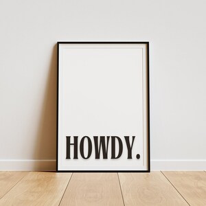 Op de afbeelding: Zwart-witte ingelijste print met het woord "HOWDY" in vetgedrukte, zwarte letters.