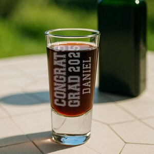 May include: A clear shot glass filled with a dark liquid, featuring the etched text "CONGRATS GRAD 2025 DANIEL." A green bottle is blurred in the background. The glass sits on a white tiled surface.