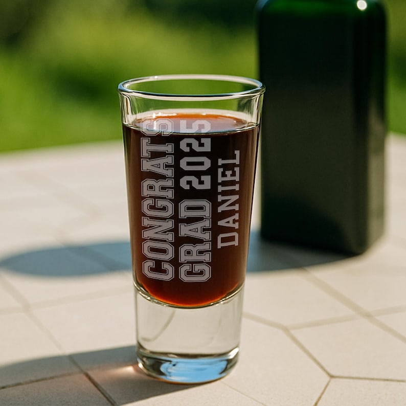 May include: A clear shot glass filled with a dark liquid, featuring the etched text "CONGRATS GRAD 2025 DANIEL." A green bottle is blurred in the background. The glass sits on a white tiled surface.
