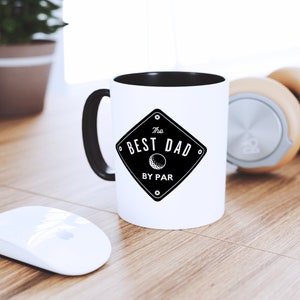May include: White coffee mug with a black handle and a black rim. The mug has a black and white graphic that says "The Best Dad By Par" with a golf ball icon.
