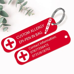 Op de afbeelding: Twee rode rechthoekige sleutelhangers met witte tekst en een wit kruissymbool. E&eacute;n sleutelhanger leest "CUSTOM ALLERGY EPI-PEN IN BAG" en de andere heeft contactgegevens. Een zilveren sleutelring verbindt de sleutelhangers.
