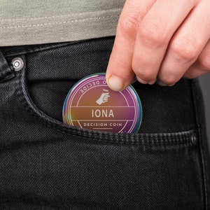 Puede incluir: Una moneda de decisi&oacute;n redonda con un degradado de colores, incluyendo morado, naranja y amarillo, se est&aacute; colocando en un bolsillo de un jean negro. La moneda tiene el nombre "IONA" y las palabras "DECISION COIN".