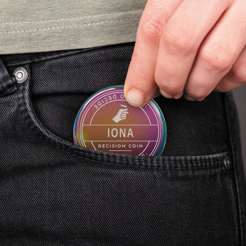 Puede incluir: Una moneda de decisi&oacute;n redonda con un degradado de colores, incluyendo morado, naranja y amarillo, se est&aacute; colocando en un bolsillo de un jean negro. La moneda tiene el nombre "IONA" y las palabras "DECISION COIN".