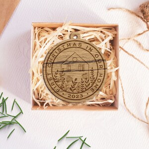 May include: A round wooden ornament with the text "First Christmas in our home 2023" and a stylized illustration of a house with a window and a door, surrounded by plants.
