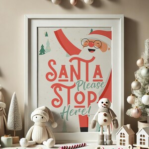 Puede incluir: Una impresi&oacute;n enmarcada en blanco con una ilustraci&oacute;n de dibujos animados de Santa Claus con el texto "Santa Please Stop Here!" en letras rojas y verdes. La impresi&oacute;n est&aacute; rodeada de un marco blanco y est&aacute; sentada sobre una mesa con una peque&ntilde;a figura de mu&ntilde;eco de nieve blanco y un peque&ntilde;o &aacute;rbol de Navidad blanco.