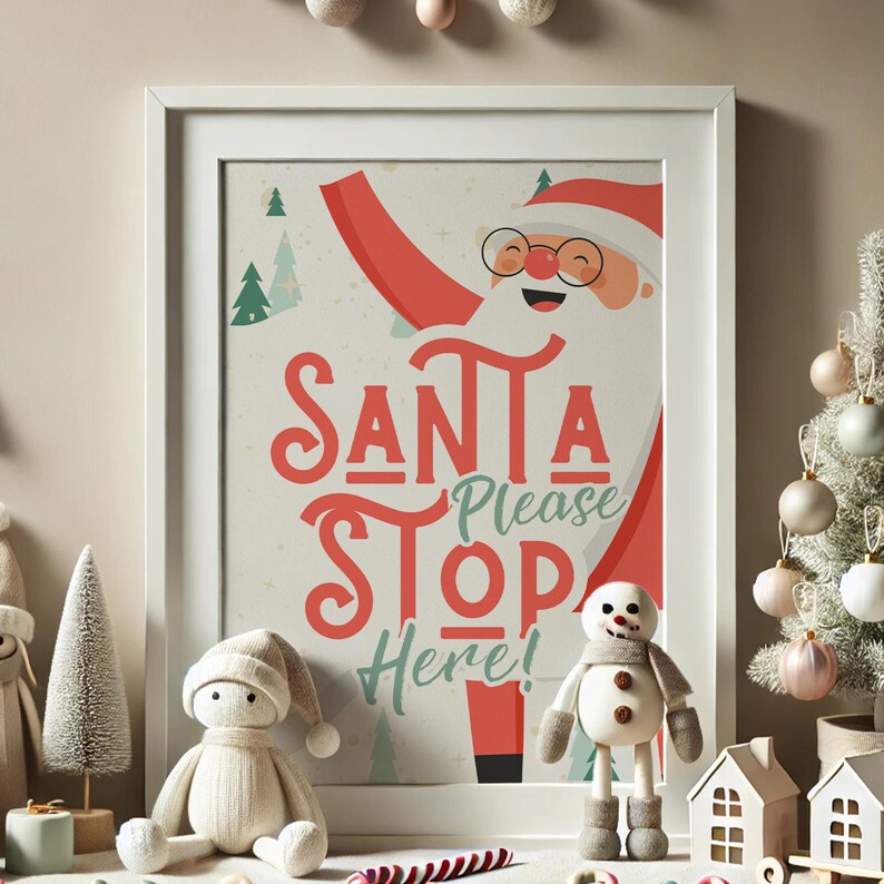 Puede incluir: Una impresi&oacute;n enmarcada en blanco con una ilustraci&oacute;n de dibujos animados de Santa Claus con el texto "Santa Please Stop Here!" en letras rojas y verdes. La impresi&oacute;n est&aacute; rodeada de un marco blanco y est&aacute; sentada sobre una mesa con una peque&ntilde;a figura de mu&ntilde;eco de nieve blanco y un peque&ntilde;o &aacute;rbol de Navidad blanco.