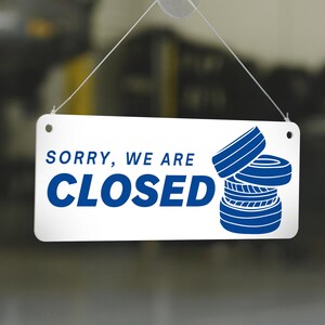 May include: White rectangular sign with the words "SORRY, WE ARE CLOSED" in blue. The sign also features a blue graphic of stacked tires. The sign is hanging by a clear string.