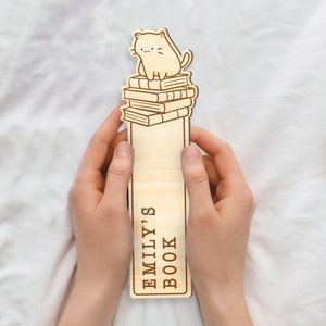 May include: Wooden bookmark with a cat sitting on top of a stack of books. The bookmark has the text "Emily's Book" on it.
