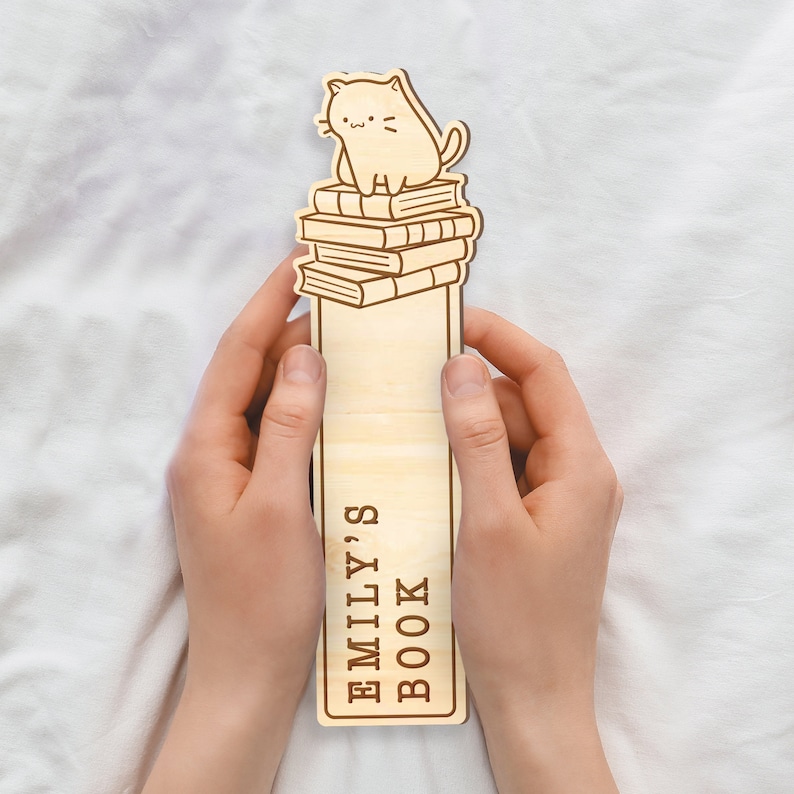 May include: Wooden bookmark with a cat sitting on top of a stack of books. The bookmark has the text "Emily's Book" on it.