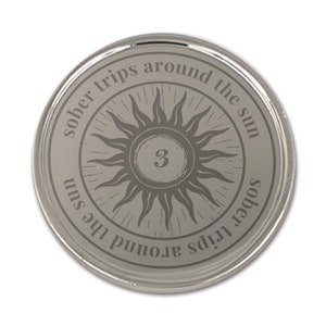 Op de afbeelding: Een zilverkleurige ronde munt met de tekst "sober trips around the sun" rond een zonnesterontwerp. Het nummer "3" staat in het midden. De munt heeft een glanzend, reflecterend oppervlak.