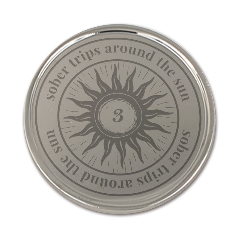 Op de afbeelding: Een zilverkleurige ronde munt met de tekst "sober trips around the sun" rond een zonnesterontwerp. Het nummer "3" staat in het midden. De munt heeft een glanzend, reflecterend oppervlak.