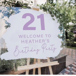 May include: A white acrylic sign with a purple paintbrush stroke design. The sign says "Welcome to Heather's 21st Birthday Party".