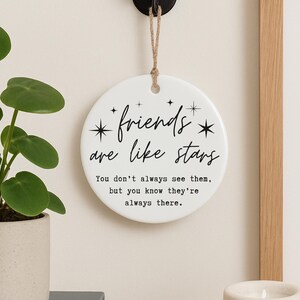 May include: A white, round ceramic wall hanging with the text "friends are like stars" in black script. The text below reads "You don't always see them, but you know they're always there." A decorative item for home decor.