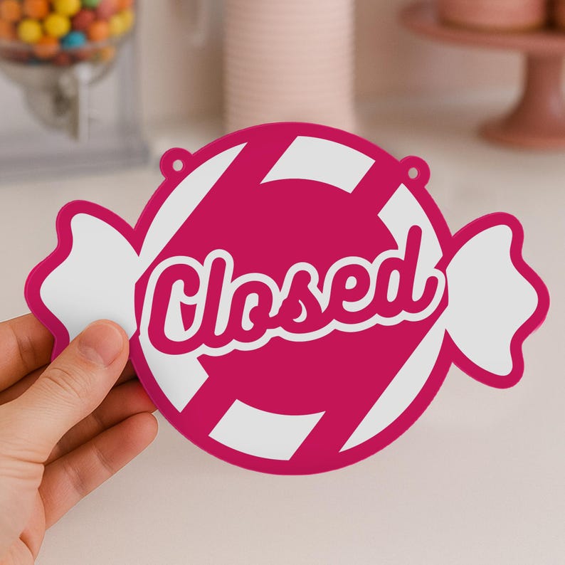 May include: A pink and white candy-shaped sign with the word "Closed" in white script. The sign has a candy wrapper design with white stripes and white wrapper ends. It is designed to be hung.