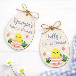 May include: Two wooden Easter egg-shaped ornaments with twine bows. One reads "Georgia's Easter Basket," the other "Holly's Easter Basket." Each features a yellow chick hatching from a pink egg, surrounded by flowers and Easter eggs.