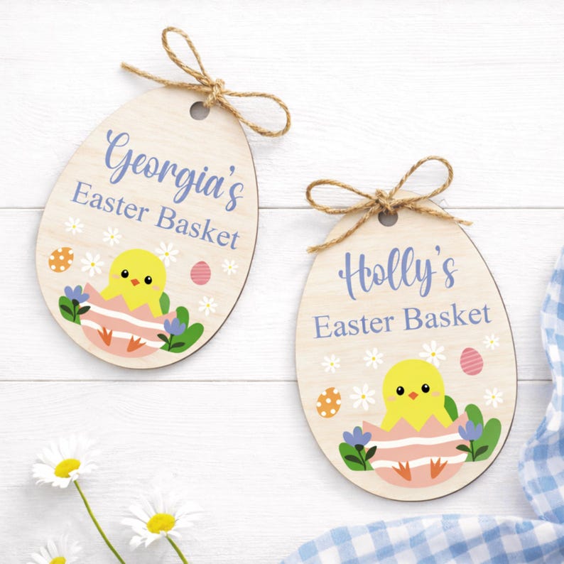 May include: Two wooden Easter egg-shaped ornaments with twine bows. One reads "Georgia's Easter Basket," the other "Holly's Easter Basket." Each features a yellow chick hatching from a pink egg, surrounded by flowers and Easter eggs.
