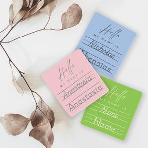 May include: Three square name tags with a cursive font. The tags are pink, blue, and green. The text on the tags reads "Hello, My Name Is" followed by a line for writing a name. The names "Anastasia", "Nicholas", and "Name" are written on the tags.
