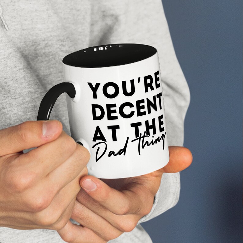 May include: White coffee mug with a black handle and black rim. The mug has black text that reads "You're Decent at the Dad Thing."