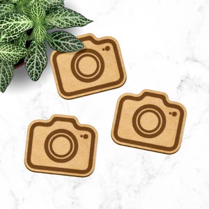 May include: Three wooden camera-shaped coasters. The coasters are light brown with a darker brown outline and a dark brown circle in the center to represent the lens.